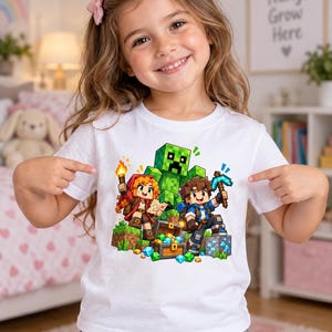 May include: A white short-sleeved t-shirt featuring a colorful graphic print of two pixelated characters, a green creeper, a treasure chest filled with gems, and a diamond pickaxe, all rendered in a blocky, video game-inspired art style.