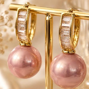Peut inclure: Une paire de créoles dorées présentant une rangée de pierres transparentes taille baguette sur le devant de l'anneau et une grosse perle ronde, lisse et rose poudré suspendue à la base de chaque boucle, créant un design pendant.
