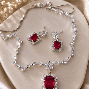 以下が含まれることがあります： ネックレスとイヤリングのジュエリーセット。ネックレスには、透明なクリスタルで囲まれた大きな長方形の赤い石のペンダントがあり、透明なクリスタルのチェーンにつながっています。イヤリングも同様のデザインです。