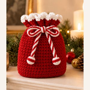 Pode incluir: Uma bolsa de crochê vermelha com cordão, borda superior festonada branca e um cordão listrado de vermelho e branco amarrado em um laço. O cordão termina em dois pequenos pompons redondos listrados de vermelho e branco, criando um recipiente.