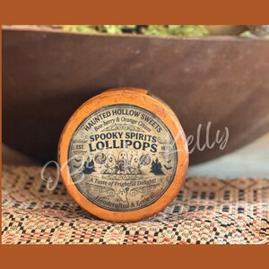 May include: A round orange container featuring a vintage-style label that reads Haunted Hollow Sweets, Boo-berry & Orange Cream, Spooky Spirits Lollipops, Est. 1873, A Taste of Frightful Delight!, and Handcrafted & Eerie with ghost illustrations.