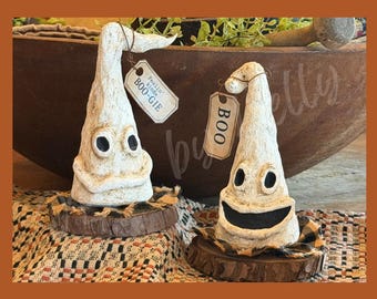 May include: Two off-white, textured ghost figurines with black eyes and mouths, each mounted on a circular wood slice base. One ghost features a tag reading "Feelin' kinda BOO-GIE" and the other has a tag reading "BOO" attached with twine.