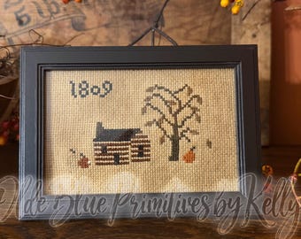 May include: A framed cross-stitch embroidery on tan fabric featuring a dark brown log cabin, a bare tree, and a small orange pumpkin. The year 1809 is stitched in the upper left corner. The piece is enclosed in a simple black rectangular frame.