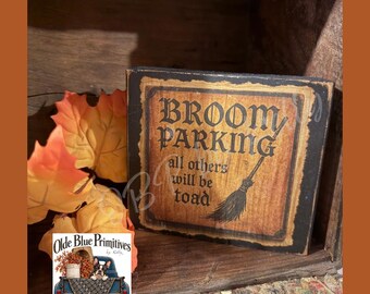 May include: A rectangular wooden sign with a distressed black border and a tan center. The text reads BROOM PARKING all others will be toad, accompanied by a black graphic of a broomstick. The sign has a weathered, aged appearance.