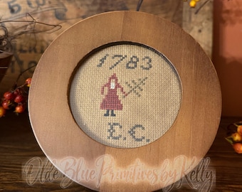 May include: A round wooden frame containing a cross-stitch sampler on beige fabric. The embroidery features the year 1783, the initials E.C., and a small figure in a red dress holding a tree. The text Olde Blue Primitives by Kelly is visible.