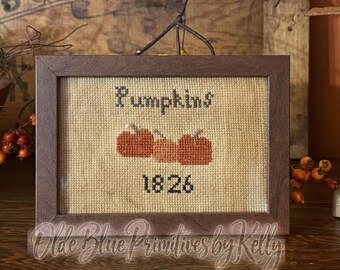 May include: A framed cross-stitch embroidery on tan fabric featuring the word Pumpkins in black, three orange pumpkins in the center, and the year 1826 in black at the bottom. The piece is enclosed in a simple dark brown wooden frame.