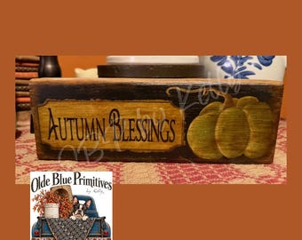 May include: A rectangular wooden block sign with a dark stained finish, featuring a central gold-toned label with the black text Autumn Blessings and a painted green pumpkin. A small logo in the corner shows a blue truck with a dog.