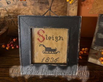 May include: A square black distressed wooden frame containing a cross-stitch sampler on tan fabric. The embroidery features the word Sleigh in red and black, a black silhouette of a sleigh, and the year 1836 stitched in black thread.