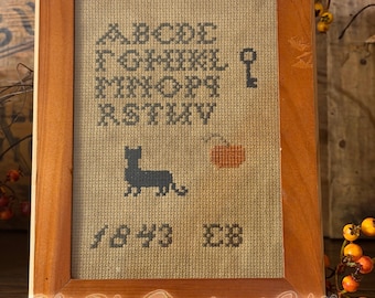 May include: A framed cross-stitch sampler on tan fabric featuring the alphabet from A to V in black thread, a black key, a black cat silhouette, an orange pumpkin, and the text 1843 EB. The piece is enclosed in a simple light brown wooden frame.