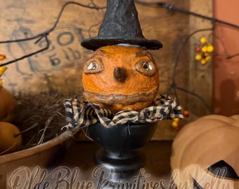 May include: A textured orange pumpkin figure with a pointed nose and painted eyes, wearing a tall black witch hat and a black and beige checkered fabric collar, mounted on a black pedestal base with the text Olde Blue Primitives by Kelly.