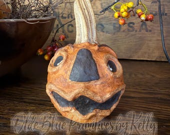 May include: A carved orange pumpkin figure with a textured surface, a tall stem, and dark, recessed facial features including eyes, a nose, and a wide mouth. The text "Olde Blue Primitives by Kelly" is overlaid at the bottom of the image.