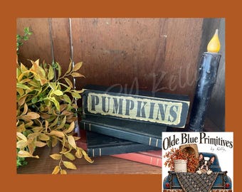 May include: A black wooden block sign with the word PUMPKINS printed in light green, stacked on two dark books. Beside the books is a black taper candle with a yellow flame, and to the left is a leafy green and yellow plant.