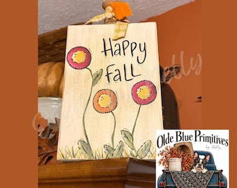 May include: A rectangular wooden sign with a cream-colored background featuring the hand-painted text Happy Fall in black. Three stylized flowers with pink petals and yellow centers grow from green stems. A fabric ribbon is tied to the top.