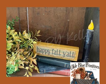 May include: A rectangular wooden block sign with a yellow finish and the text "happy fall y'all" in dark lettering. The sign rests on a stack of two dark-colored books next to a black taper candle with a yellow flame and green foliage.