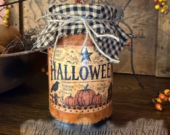 May include: A cylindrical jar with a textured orange finish, featuring a label with the text HALLOWEEN, pumpkins, and a crow. The lid is covered with a black and white checkered fabric tied with twine, creating a rustic appearance.