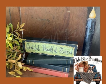 May include: A rectangular wooden sign with a green center panel featuring the black text Grateful Thankful Blessed, resting on a stack of three books in black and red, accompanied by a black taper candle and a sprig of green leaves.