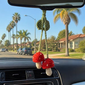 May include: A crochet car accessory hanging from a rearview mirror, featuring two red mushrooms with white spots and cream-colored stems. The mushrooms are attached to green braided cords and a small green leaf accent, creating a hanging decoration.