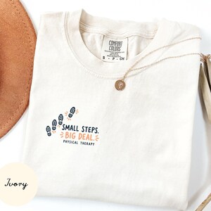 May include: An ivory t-shirt with a Comfort Colors label, featuring a graphic of four dark blue footprints and the text "SMALL STEPS. BIG DEAL. PHYSICAL THERAPY" in dark blue and orange. A gold-toned necklace with a small circular pendant rests on top.