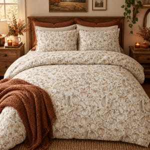 Peut inclure: Une parure de lit composée d'une housse de couette blanche et de deux taies d'oreiller assorties avec un motif répétitif de citrouilles au trait marron. Un plaid en tricot épais et texturé orange brûlé est posé sur le lit.