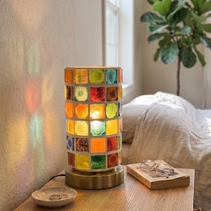 Könnte beinhalten: Eine zylindrische Tischleuchte mit einem Mosaik aus quadratischen, bunten Glasfliesen in Rot-, Orange-, Gelb-, Grün- und Blautönen. Die Lampe hat einen runden, messingfarbenen Metallsockel und strahlt ein warmes, leuchtendes Licht durch das Glas.