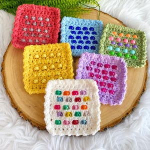 May include: Six square crochet coasters in various colours including red, light blue, green, yellow, purple, and white. Each coaster features a grid of small, translucent plastic beads sewn into the centre of the crocheted yarn fabric.