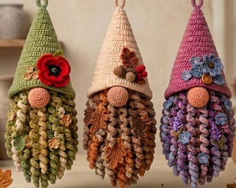 May include: Three hanging crochet gnomes with pointed hats. The left gnome has a green hat with a red poppy. The center gnome has a beige hat with acorns. The right gnome has a purple hat with blue flowers. All have curly beards and round noses.