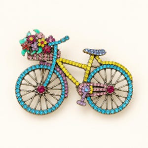 May include: A bicycle-shaped brooch featuring a metallic frame encrusted with small, multicolored rhinestones. The bike includes a front basket filled with pink, red, yellow, and purple floral accents, with blue, yellow, and pink stones throughout.