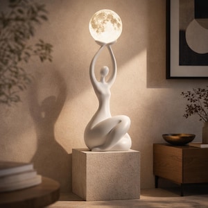 Peut inclure: Une lampe de table sculpturale blanche représentant une silhouette humaine stylisée en position assise, les bras levés au-dessus de la tête, soutenant un globe sphérique texturé rappelant la lune, diffusant une lumière chaude et douce.