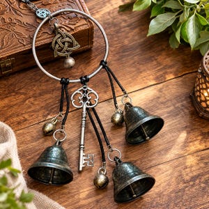 May include: A decorative wall hanging featuring a large silver-toned metal ring adorned with a bronze-colored triquetra charm, a silver-toned skeleton key, and four bells of varying sizes. Black leather cords attach the charms to the ring.