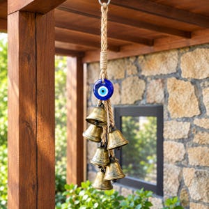 May include: A hanging wind chime featuring a thick, braided jute rope cord topped with a metal ring. A circular blue glass evil eye bead sits above a cluster of five antique-style brass bells of varying sizes, all suspended from the central rope.