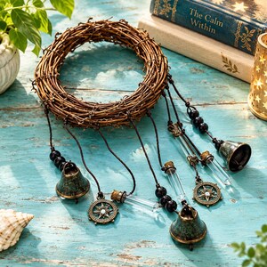 May include: A circular brown twig wreath adorned with hanging elements including two weathered green metal bells, two clear glass vials with cork stoppers, and two circular bronze-toned compass charms, all suspended by dark brown cords with black beads.
