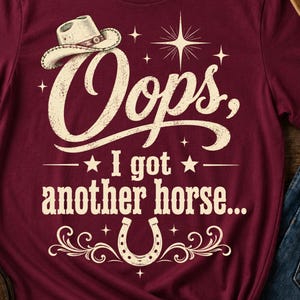 Peut inclure: Un t-shirt à manches courtes bordeaux avec une impression graphique crème. Le motif comprend un chapeau de cow-boy, un fer à cheval, des étoiles et le texte : Oops, I got another horse... L'écriture est dans une police vieillie.