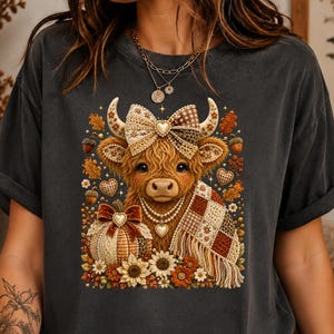 Peut inclure: Un t-shirt gris anthracite avec un motif de vache Highland portant un nœud à carreaux et un collier de perles. La vache est drapée dans une couverture à carreaux à côté d'une citrouille, entourée de feuilles d'automne et de tournesols.