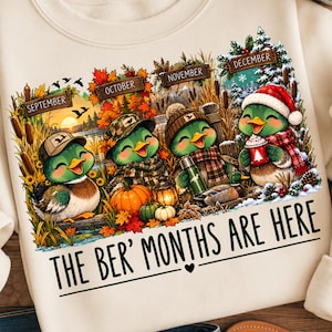 Peut inclure: Un sweat-shirt couleur crème orné d'un motif représentant quatre canards de dessin animé pour septembre, octobre, novembre et décembre. Le design inclut des éléments saisonniers comme des citrouilles, des feuilles d'automne et de la neige, avec le texte THE BER' MONTHS ARE HERE.