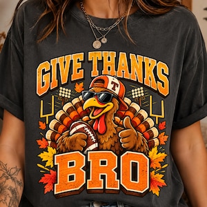 May include: A charcoal grey t-shirt featuring a graphic of a turkey wearing a baseball cap and sunglasses, holding a football and giving a thumbs up. The text reads GIVE THANKS BRO, surrounded by football goalposts, stadium lights, and autumn leaves.