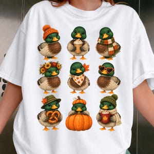 Peut inclure: Un t-shirt blanc avec une impression graphique de neuf canetons colverts portant diverses tenues et accessoires sur le thème de l'automne, notamment un bonnet en tricot, une écharpe, une couronne de tournesols, des lunettes en forme de cœur, une citrouille et un bretzel.
