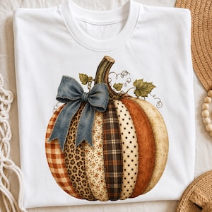 Peut inclure: Un t-shirt blanc orné d'un grand motif de citrouille composé de panneaux verticaux aux motifs variés, notamment vichy, léopard, floral, écossais et à pois. Un nœud bleu à texture denim est attaché à la tige de la citrouille.