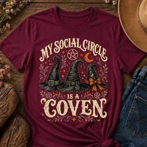 Peut inclure: Un t-shirt à manches courtes bordeaux avec une impression graphique représentant trois chapeaux de sorcière noirs, un croissant de lune et des pentagrammes. Le texte indique "MY SOCIAL CIRCLE IS A COVEN" avec des motifs floraux et des étoiles.