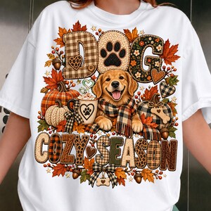 Peut inclure: Un t-shirt blanc avec un motif représentant un golden retriever portant une écharpe à carreaux, entouré de citrouilles, de feuilles d'automne, de glands et de cœurs. Le texte DOG COZY SEASON est écrit en grandes lettres à motifs.