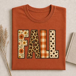Op de afbeelding: Een roestkleurig t-shirt met het woord FALL in grote blokletters. Elke letter heeft een eigen patroon: de F heeft een bloemenprint, de A een luipaardprint, de L een oranje gingham ruitje en de tweede L heeft bruine stippen.