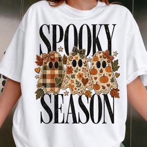 Peut inclure: Un t-shirt blanc avec le texte noir SPOOKY SEASON et trois illustrations de fantômes. Les fantômes portent des nœuds et sont décorés de motifs à carreaux, floraux et de citrouilles, entourés de petites feuilles d'automne, d'étoiles et de cœurs.