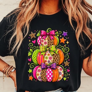 Peut inclure: Un t-shirt noir avec un motif de trois citrouilles empilées décorées de motifs colorés, notamment des imprimés léopard, des pois et des fleurs. Les citrouilles sont ornées de nœuds et d'étoiles aux couleurs rose vif, vert lime et violet.