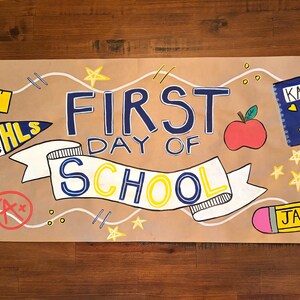 Puede incluir: Una pancarta rectangular de papel marr&oacute;n con el texto FIRST DAY OF SCHOOL dibujado a mano en azul y amarillo. Las ilustraciones incluyen una regla amarilla etiquetada CHARLIE, una mochila azul con WILL, un cuaderno azul con KATE y un l&aacute;piz amarillo con JACOB.