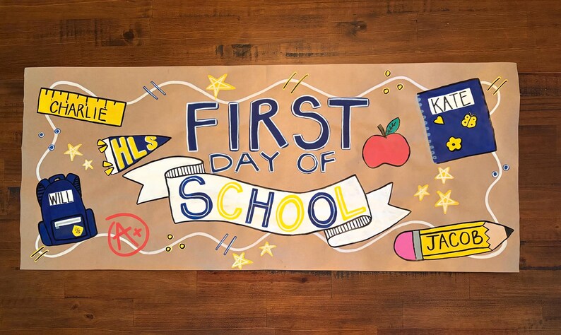 Puede incluir: Una pancarta rectangular de papel marr&oacute;n con el texto FIRST DAY OF SCHOOL dibujado a mano en azul y amarillo. Las ilustraciones incluyen una regla amarilla etiquetada CHARLIE, una mochila azul con WILL, un cuaderno azul con KATE y un l&aacute;piz amarillo con JACOB.