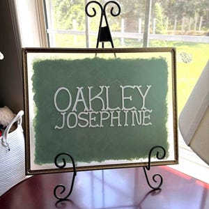 Puede incluir: Un letrero rectangular de cristal en un marco fino de metal dorado, con un fondo pintado en verde salvia y el texto blanco OAKLEY JOSEPHINE en una fuente decorativa, sobre un soporte de caballete de metal negro.