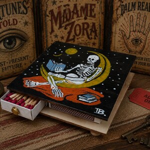 May include: A wooden matchbox drawer featuring a hand-painted black lid with a white skeleton sitting on a yellow crescent moon, reading a book against a starry night sky. The drawer contains wooden matches with red tips and a gold knob handle.