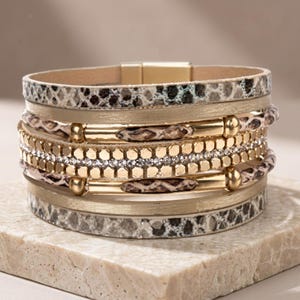 Könnte beinhalten: Ein mehrreihiges Wickelarmband mit abwechselnden Schichten aus Kunstleder mit Schlangenmuster, glatten goldfarbenen Metallbändern und einer mittleren Reihe aus kleinen, klaren Strasssteinen, flankiert von goldenen sechseckigen Perlen und Metallakzenten.