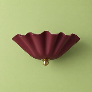 May include: A wall-mounted light fitting featuring a deep burgundy, matte-finished, ruffled shade with a wide, flared opening. The centre of the shade is accented with a small, polished brass spherical knob that serves as a decorative detail.