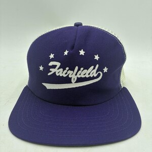 Puede incluir: Una gorra tipo camionero de color morado con un logotipo bordado en blanco en letra cursiva que dice Fairfield, acentuado por cinco pequeñas estrellas blancas dispuestas en arco sobre el texto. Tiene un panel frontal morado y malla blanca.
