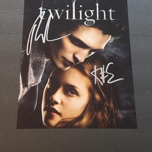 Op de afbeelding: Een rechthoekige promotieposter voor de film Twilight met een donker, sfeervol portret van twee personen met de titeltekst Twilight bovenaan. De afbeelding bevat twee duidelijke handtekeningen in witte inkt over de gezichten heen.